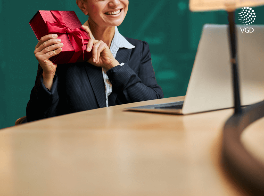 business woman trzyma w dłoniach czerwony prezent i pokazuje go przed laptopem do kamery uśmiechając się
