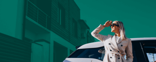 Kobieta stoi obok samochodu elektrycznego, patrzy w dal, zasłaniając oczy ręką od słońca. W tle jest segment.